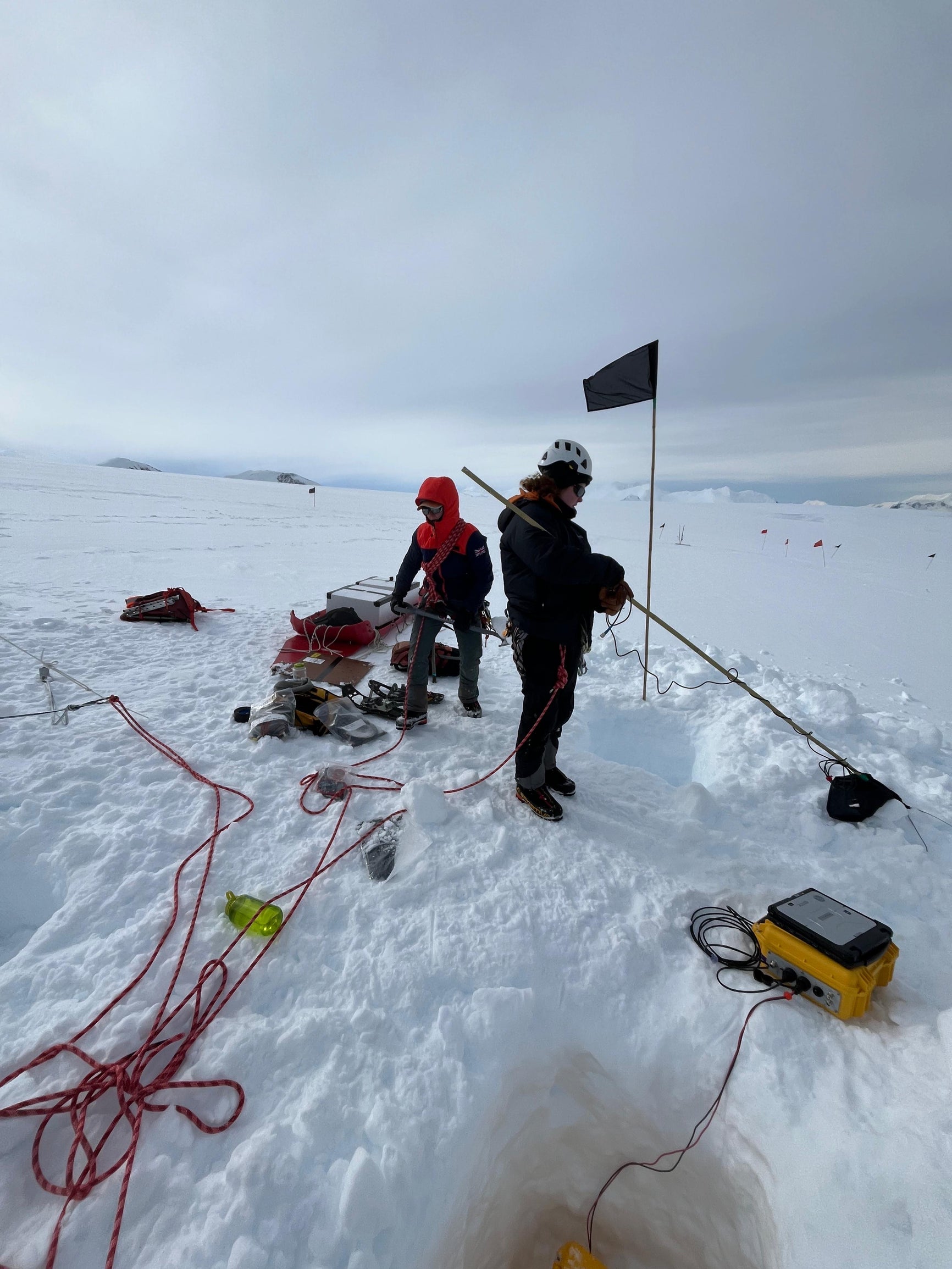 Image of 2 people next to a black flag that mark experimental radar instrument deployments at Rothera Station in Antarctica. 