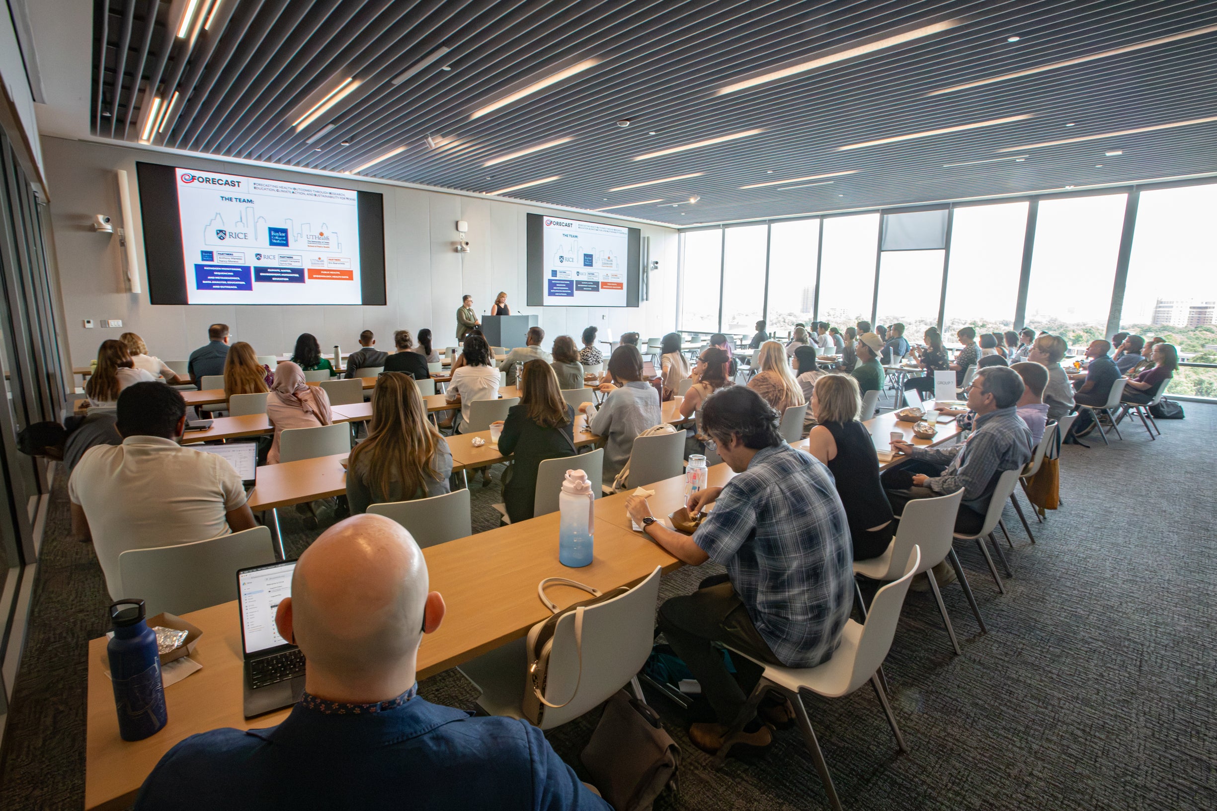 Photo of seated guests in a conference room with two speakers standing at a podium between two projector screens