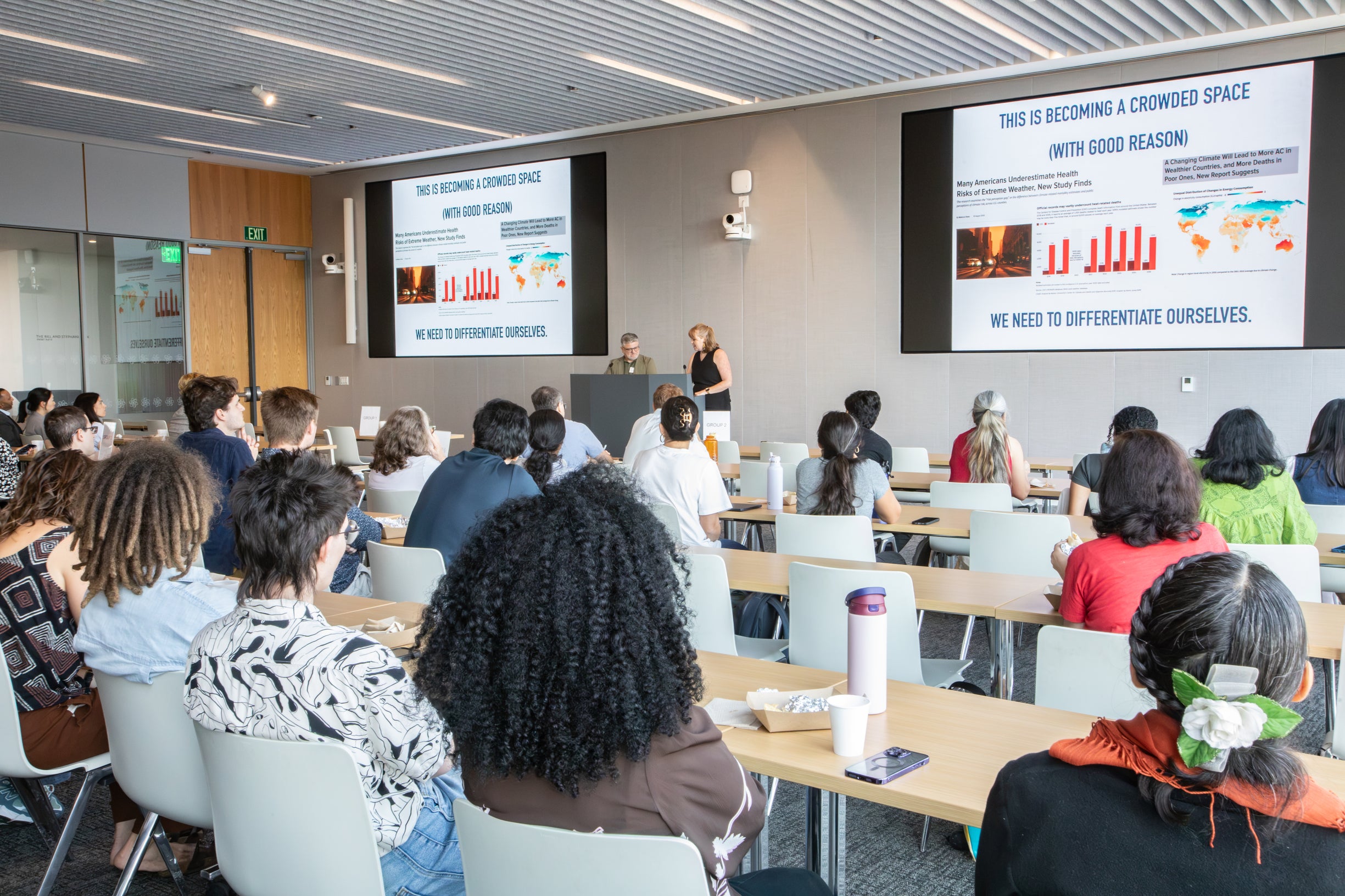 Photo of a conference room with many seated participants, two speakers standing between two projection screens