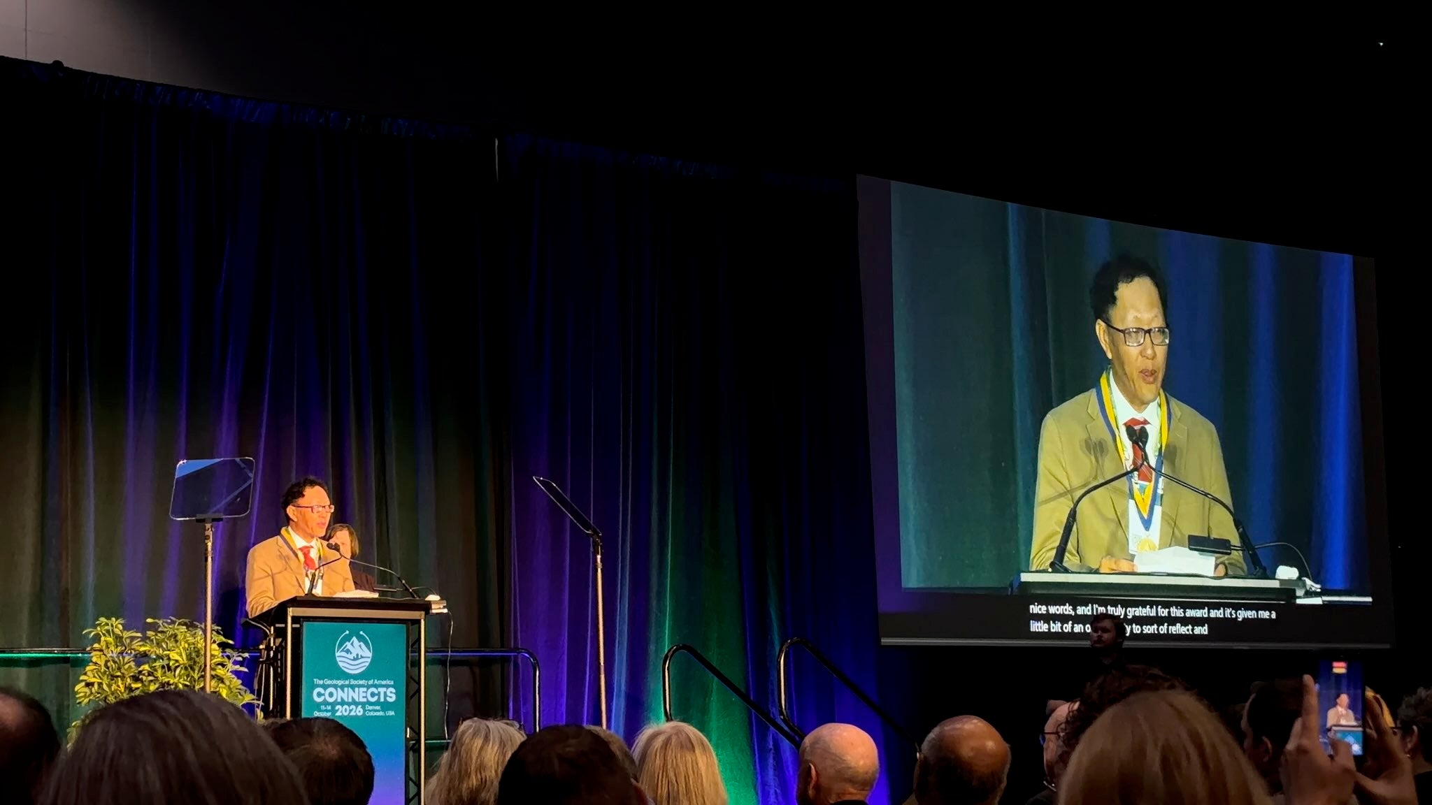 Photograph of man behind podium talking with large projection screen to the right and audience in the foreground
